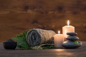 This is a decorative image of two lit, white, pillar candles set among items such as a rolled towel, balanced stones, fern leaves and more. The image indicates the relaxing and restful environment of Meagan Bombero, LMT Massage & Reiki Therapy